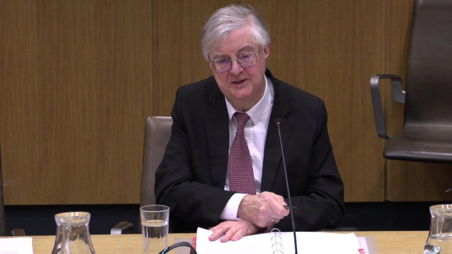 Mark Drakeford sitting at a desk in the Finance Committee, wearing glasses, a dark suit, a shirt and tie. He has a file of papers,a glass of water, a jug and a long-stemmed microphone in front of him.