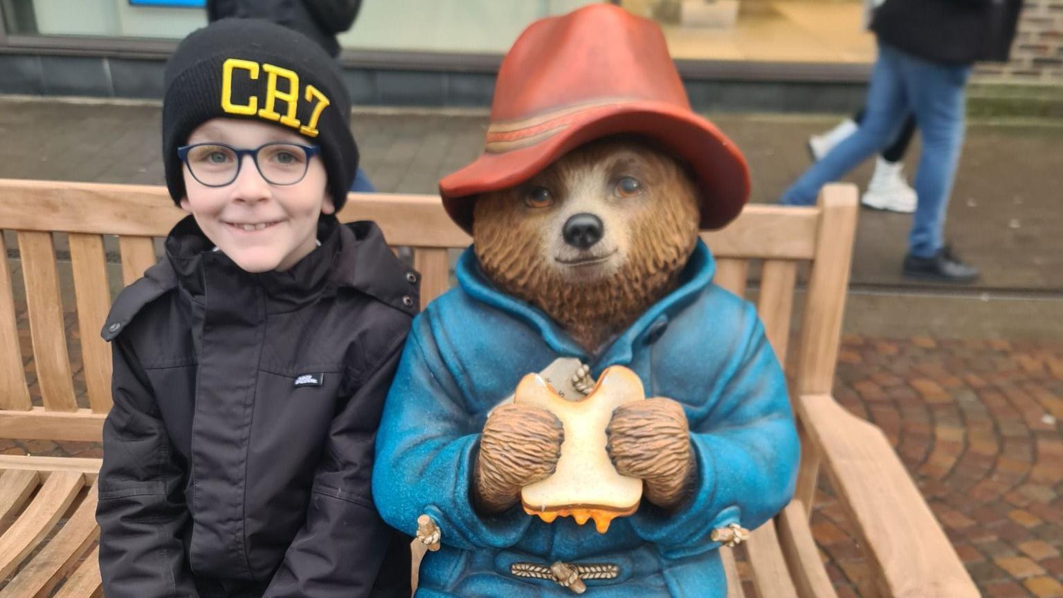 A boy is smiling on the Paddington Bear bench. People are seen walking behind them. It is a cloudy day.