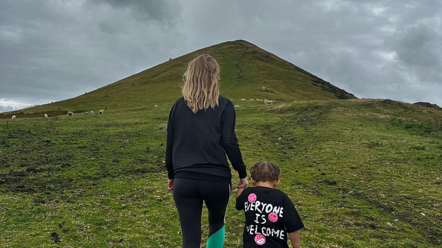 Jodi and Karter walking up a large hill. Jodie has long blonde hair past her shoulders and a black top and trousers. We see the back of both of them, Karter has a black T-shirt with the logo "Everyone is Welcome" on it