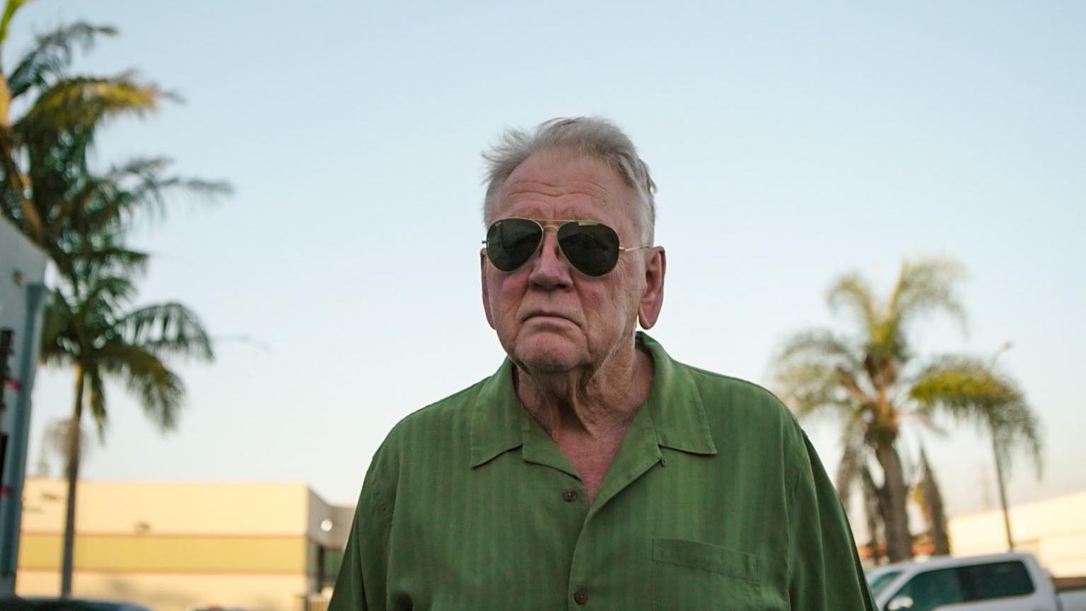 Gerry Wills who has white hear and is wearing sunglasses and green shirt. Behind him are palm trees and parked cars. 