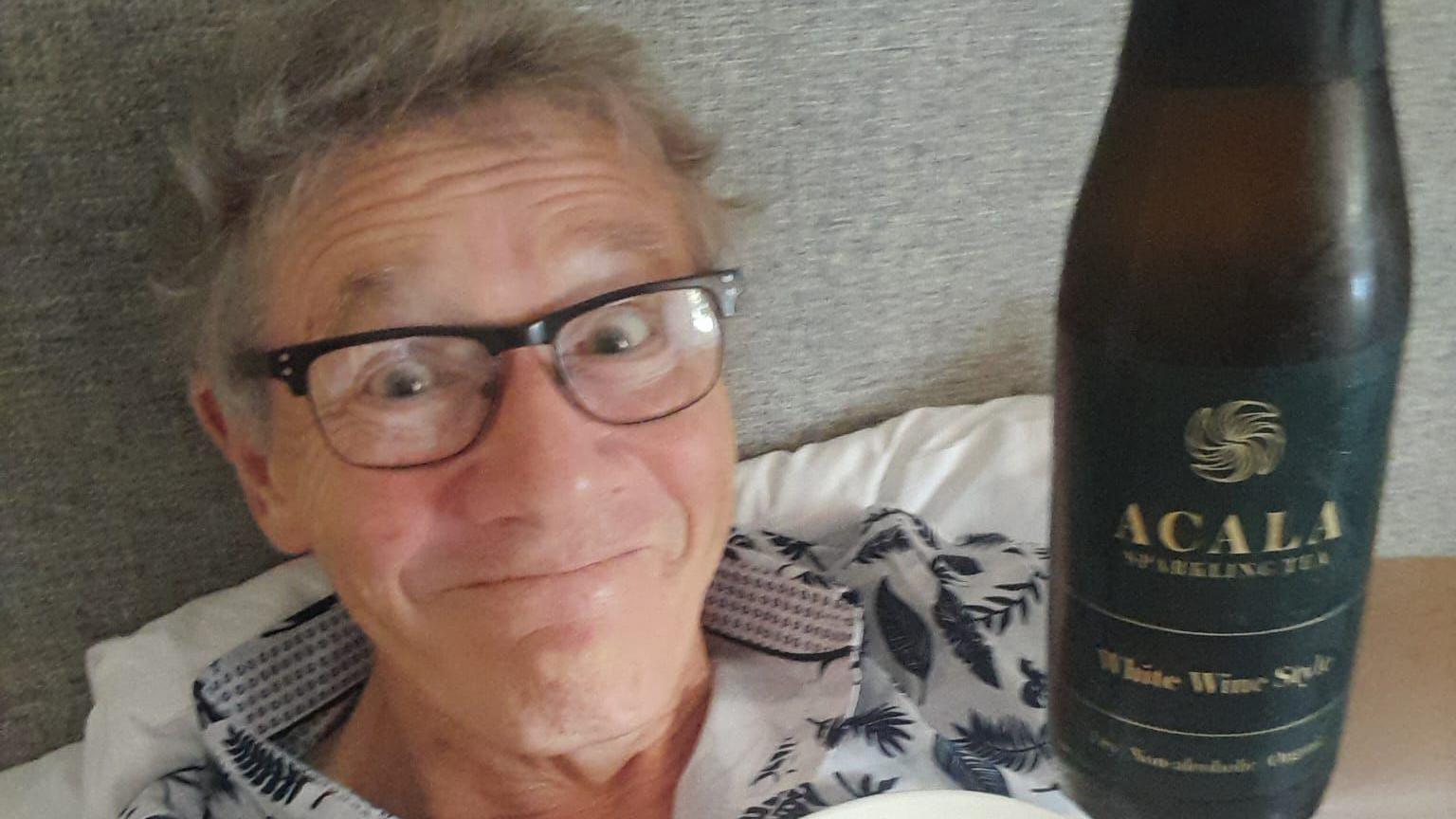 A man with grey hair and glasses lying in a bed holding up a brown bottle of sparkling "white wine style drink"