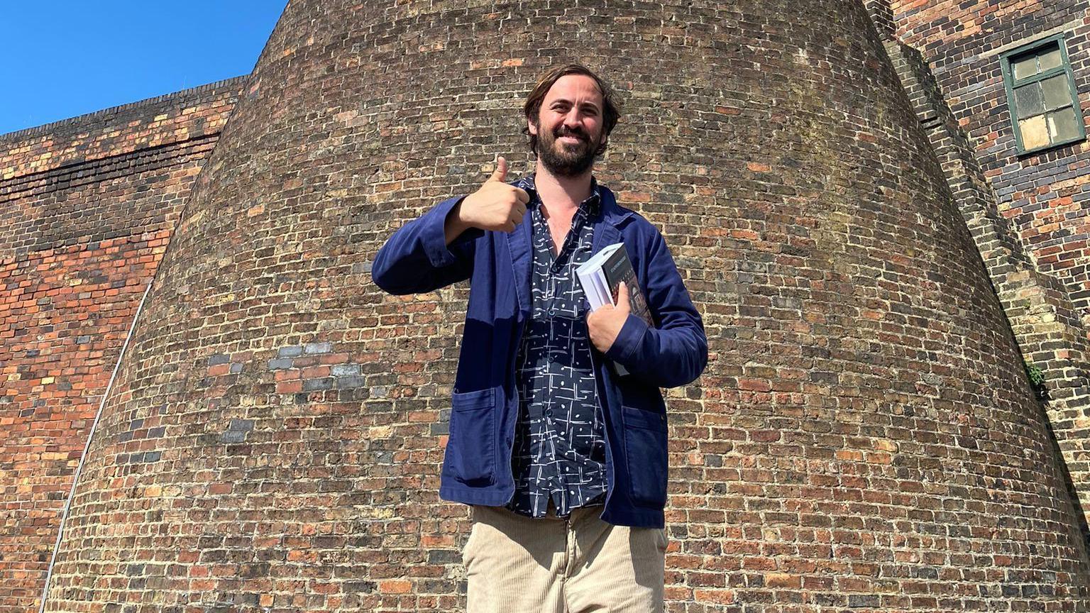 A man standing with his thumbs up in the sun, in front of a brick kiln. He has dark hair and a beard, and is wearing a blue jacket and beige trousers.