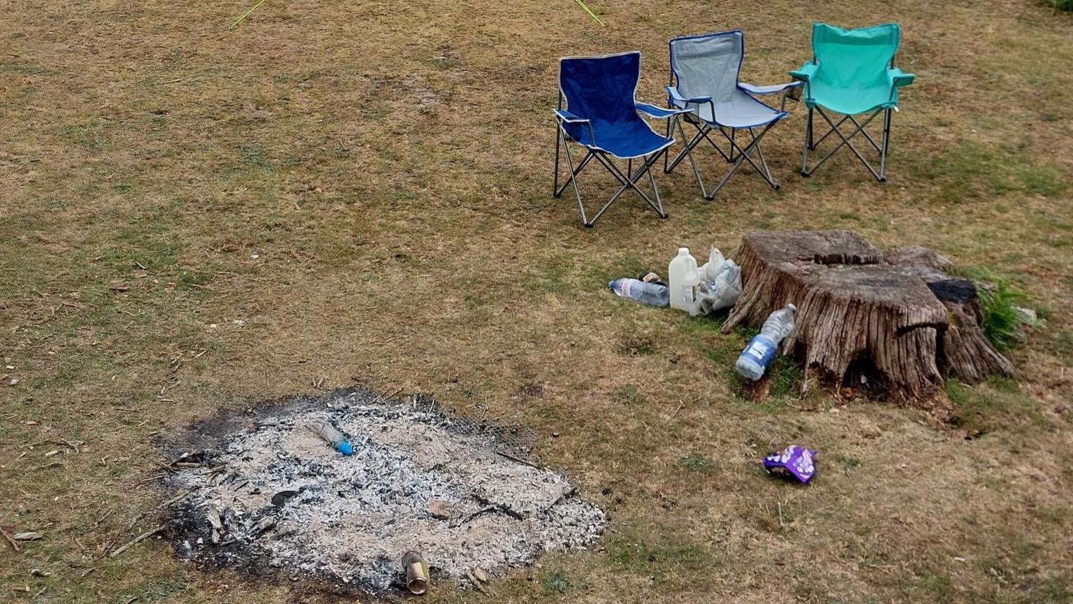 Three camp chairs, one green, one blue and one grey are next to a tree stump and the remains of a campfire. There are bottles, cans and wrappers littered on the dry looking grass.
