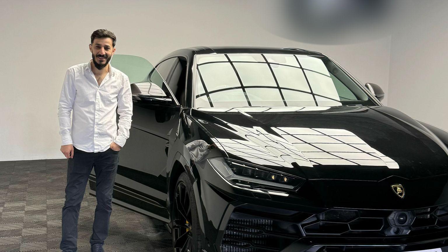 Benjamin Mustata, a man with dark hair and a beard, wearing a white shirt and dark trousers, smiles as he stands next to a black Lamborghini SUV with the driver's door open. The picture is taken indoors, in what appears to be a showroom