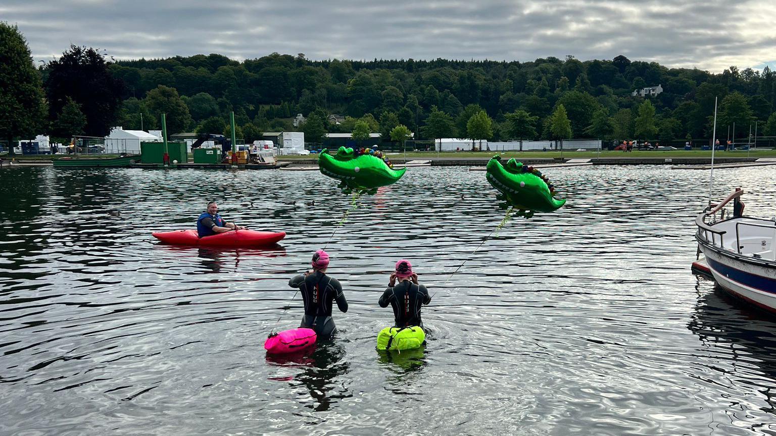 The twins are walking into the water, wearing their wetsuits and pink swim caps and have the crocodile balloons attached to them, there is a man in a red kayak on the water talking to them