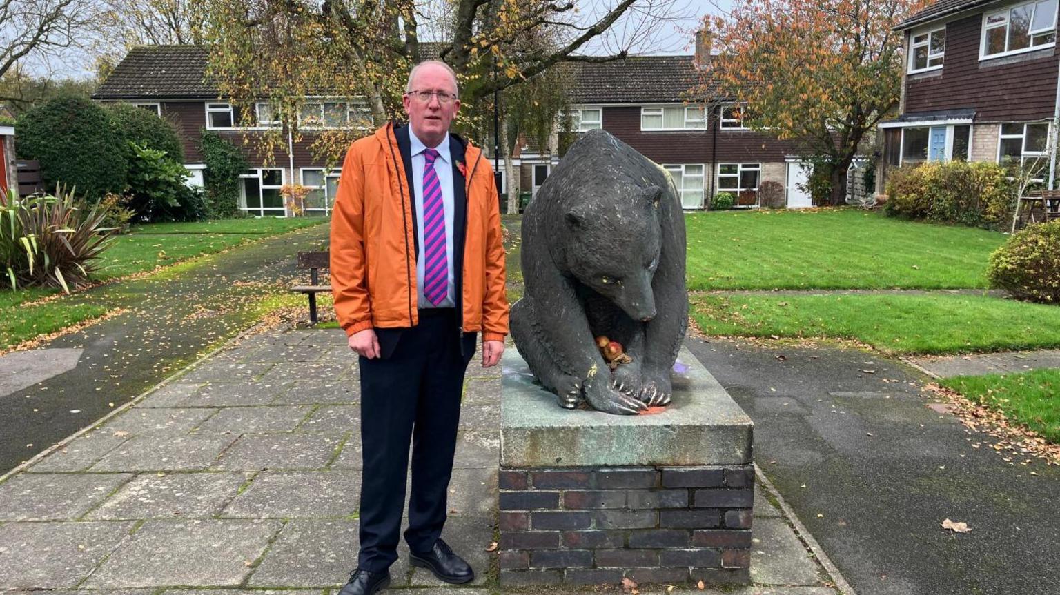 A sculpture of a bear in a square in the centre of houses. The sculpture sits on a brick plinth and there is a metal plaque attached. Next to the sculpture is a man standing. He is wearing black trousers, an orange jacket and a purple and pink striped tie. 