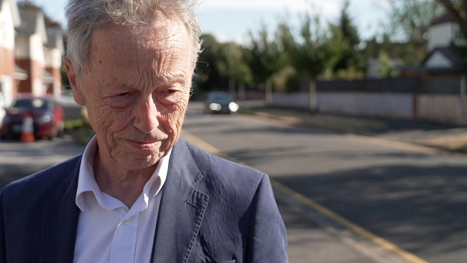Sefton Councillor Dr John Pugh with grey hair wearing a navy suit jacket and white shirt. He is standing in a residential street.