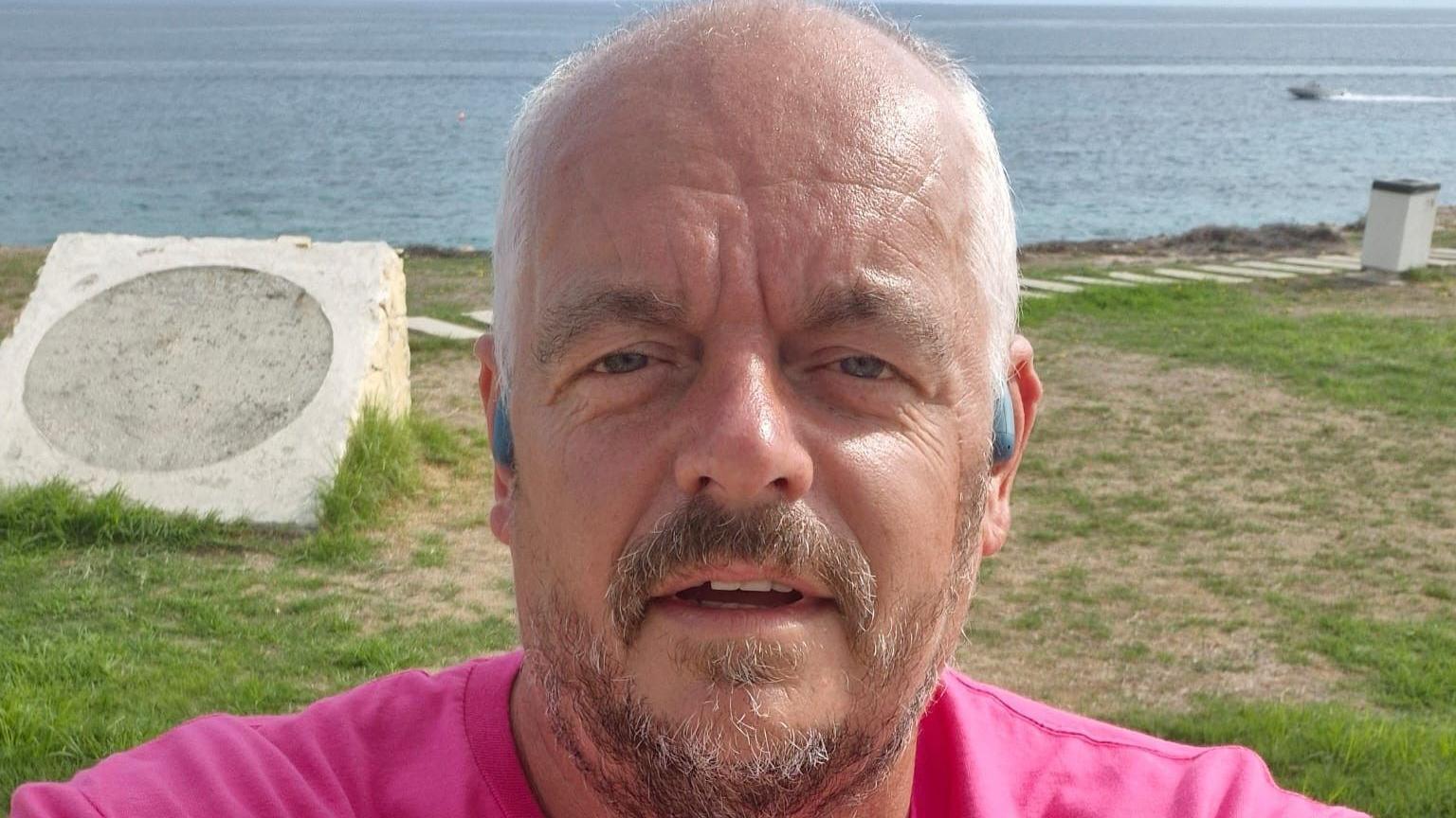 A man with very short white hair and brown and grey facial hair is taking a selfie. He is wearing grey in-ear headphones and a pink t-shirt. There is grass in the back