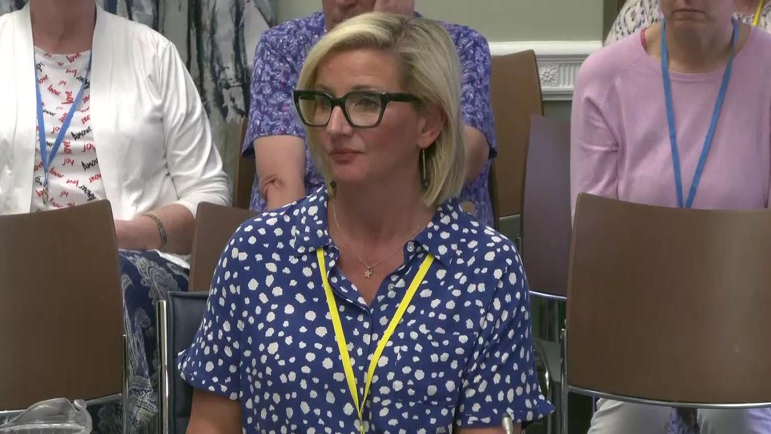 A screen grab of Roz McFeeters. She is sitting down and has short blonde hair and wearing black glasses. She has a blue top on that has white spots. She is wearing bright yellow lanyard around her neck.