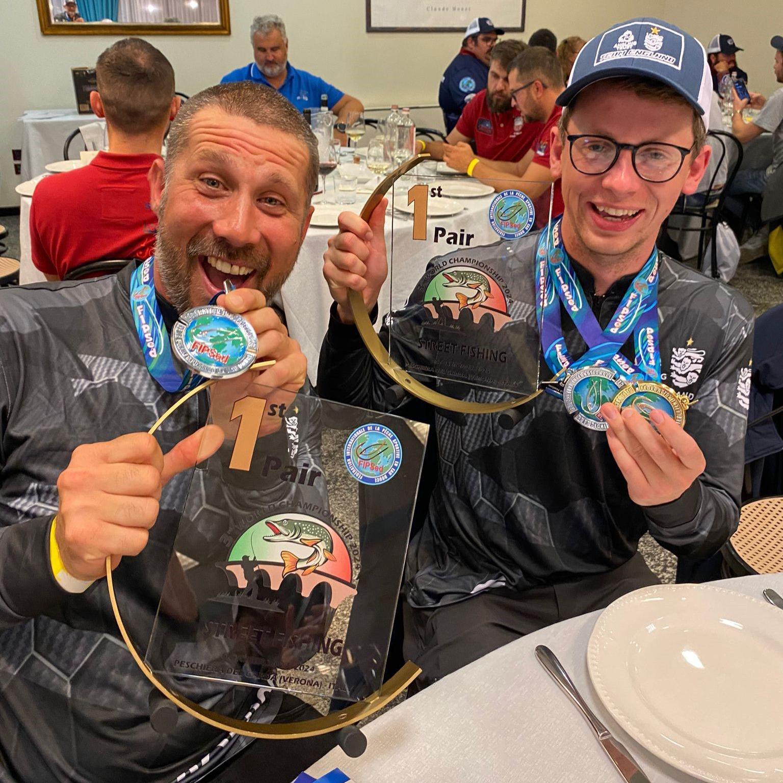 Two men hold up medals and trophies. They are sitting down and smiling at the camera.