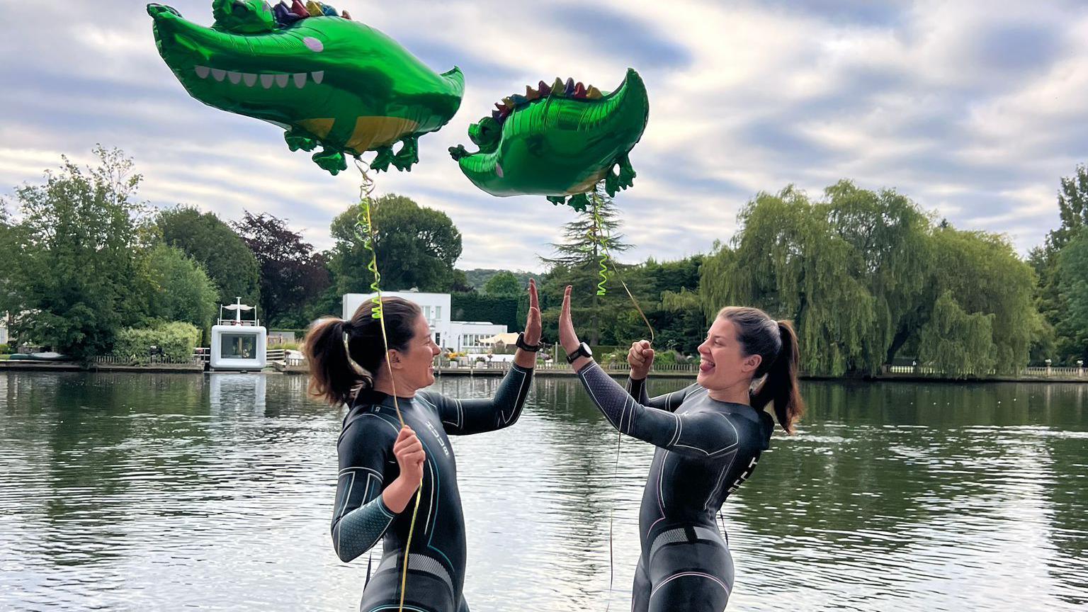 The twins stand in front of the River Thames, wearing their wetsuits and holding the two crocodile balloons high fiving each other