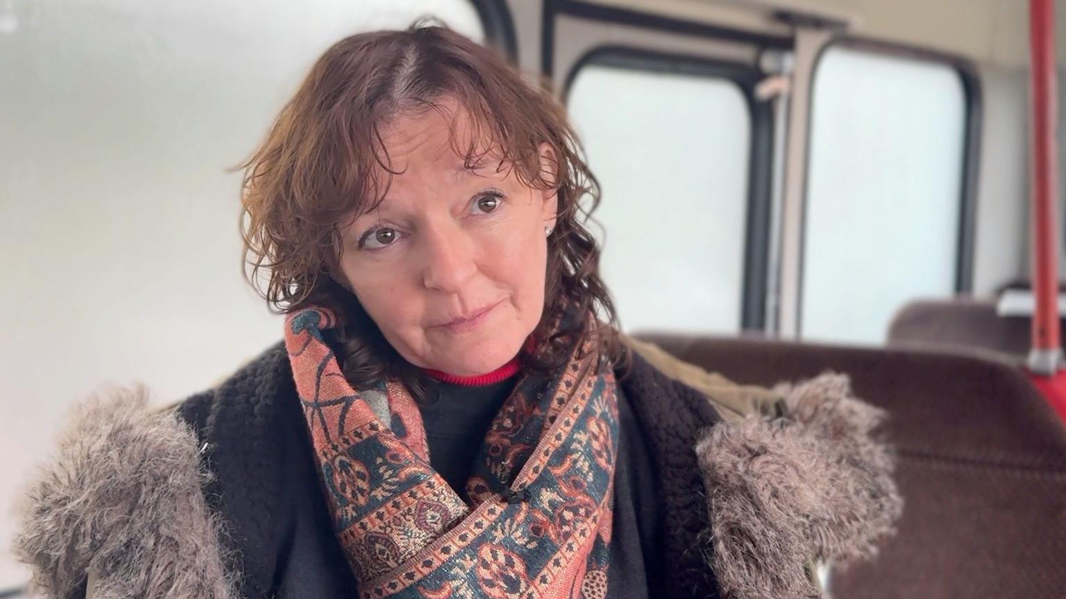 A woman in a floral scarf and jacket sits on a vintage bus. The windows are translucent and the ceiling is white. 