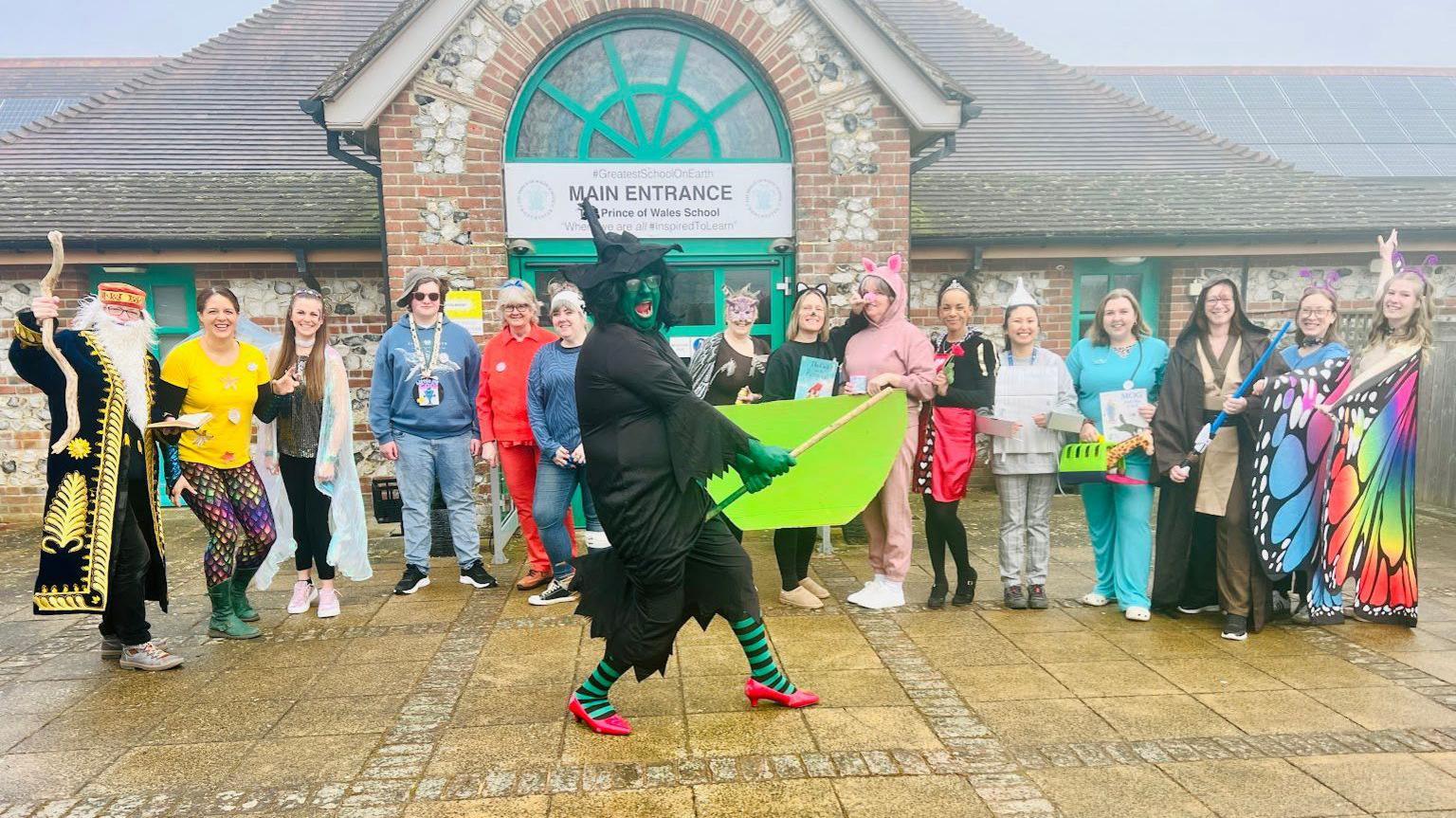 Staff in various costumes stood behind the Wicked Witch from the Wizard of Oz - who is wearing a black costumes and has their face painted green - in front of the school's entrance 