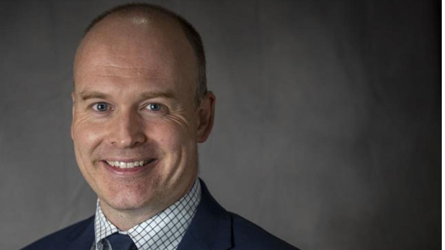 Head and shoulders shot of Andrew Crookham. He is wearing a blue suit, white and grey shirt and a blue tie. He has short hair and is smiling at the camera. He is standing in front of a grey backdrop.