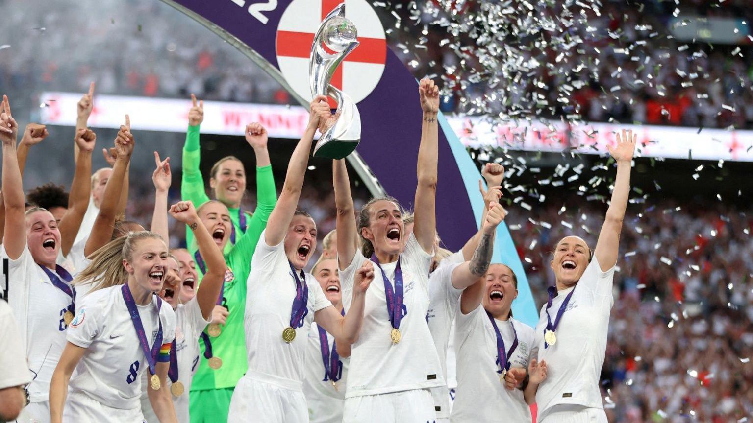 A group of female England football players lift the trophy on the pitch as confetti falls. 