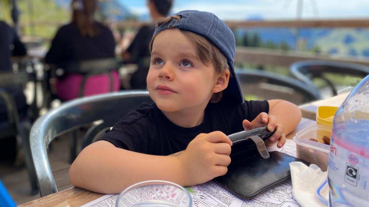 A five-year-old boy in a black shirt and blue backwards hat. He is holding a phone and looking up towards the corner of the image.