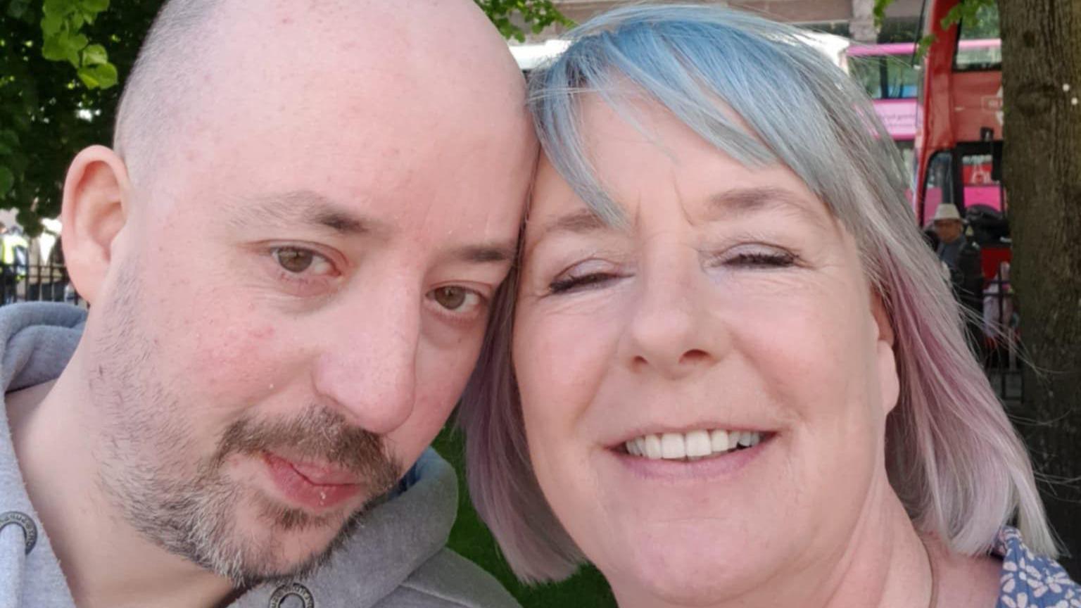 Brian is pictured in a selfie with his mother Isobel, Brian is bald and goatee and is wearing a grey hoodie. Isobel has a blue fringe and pink highlights on her short light hair. They are standing outside near buses.  