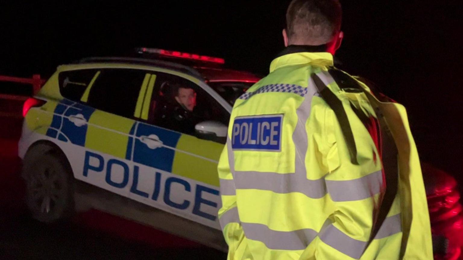 A police officer in a yellow jacket looking at an officer driving a car with yellow and blue chequered stickers and the word police, in the dark, with red lights on top of the car.