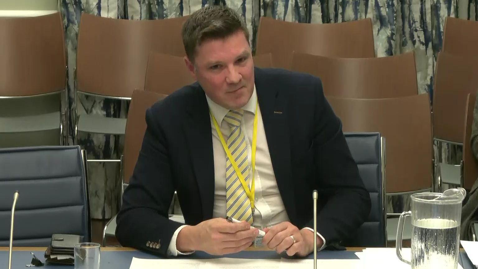 David Smyth from the Evangelical Alliance Northern Ireland giving evidence to Stormont's Education Committee.  He has short hair and is wearing a dark suit, a light shirt and a striped tie