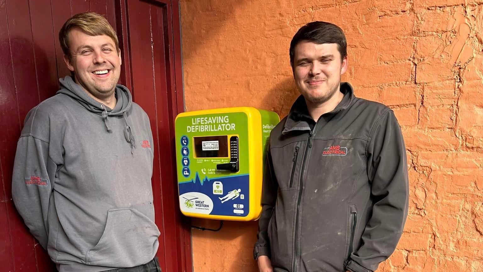 Two men smiling, standing either side of a newly-fitted defibrillator.