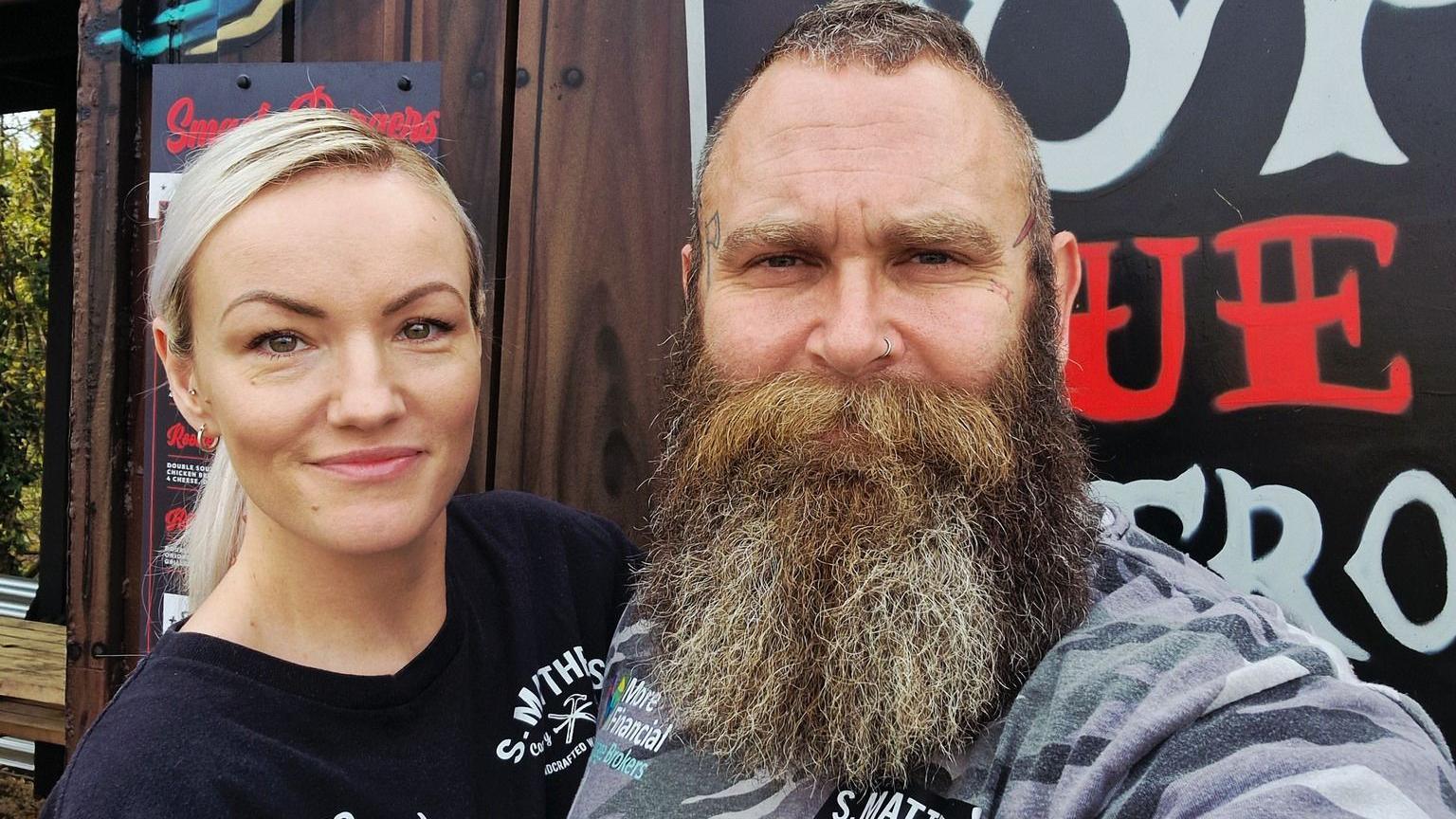 Amber, who has golden hair and is wearing a black t-shirt, stands next to Adam, who has light brown hair and long brownish-grey beard. They are standing in front of their burger shed.