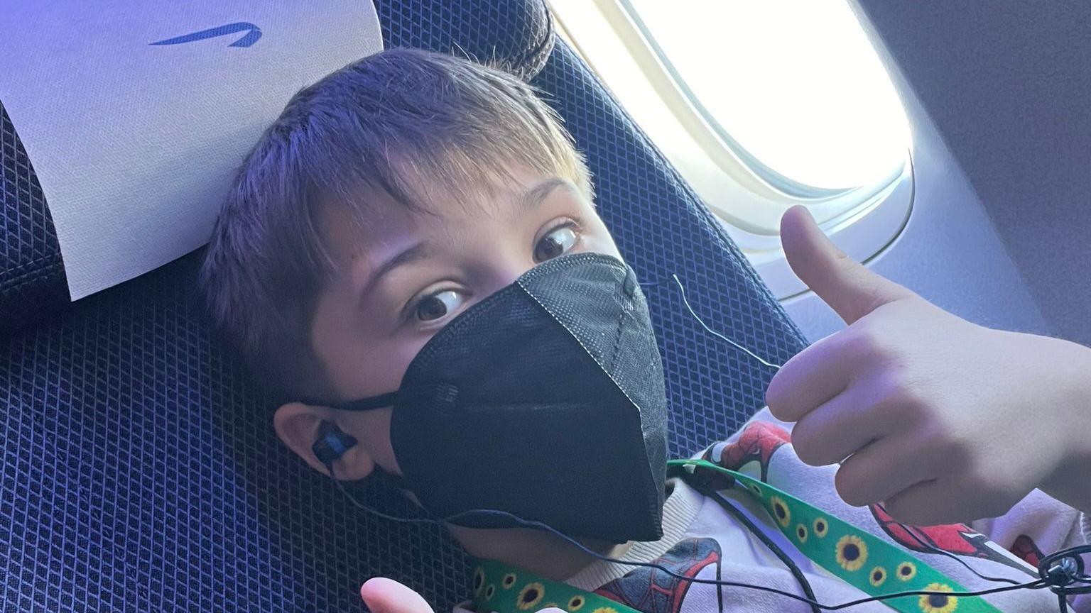 Yann Jennings, a 10-year-old boy, sat in an airplane seat wearing a black face mask, headphones, and a green lanyard with sunflowers on it. Giving thumbs up to the camera.   