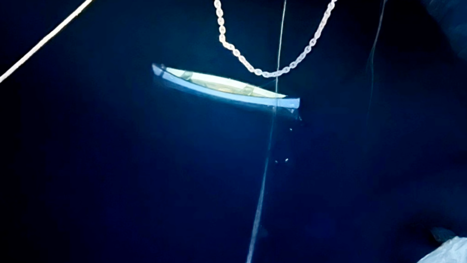 A small boat floating on dark water. It is a blue canoe - there appears to be water in it as well. Ropes and chains are visible. The lighting is dim, which creates a strong contrast between the illuminated boat and the dark surroundings, giving the scene a moody, atmospheric feel