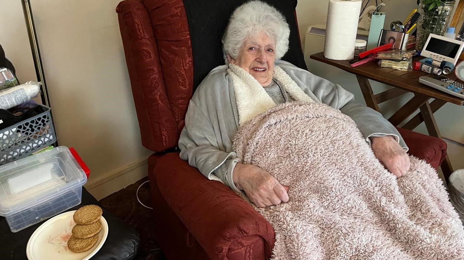 Margaret Linehan is seen with white, curly hair siting in a red armchair, smiling gently. She is wrapped in a blanket and wears a light grey cardigan over a darker top. To her left, a black leather footstool holds a plate with three biscuits and a plastic container. 