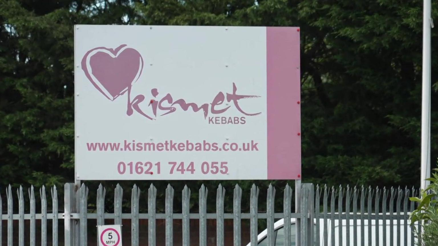 The white and red external sign of Kismet Kebabs in front of a hedge and above a metal fence