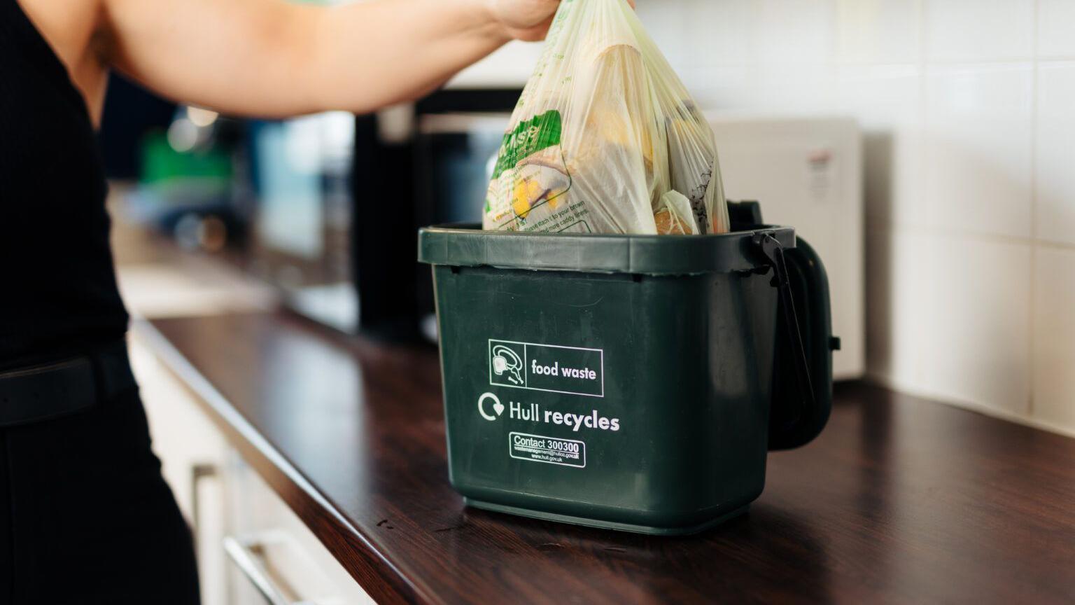 A person is placing a green food recycling bag, full of food waste, into a green Hull city council food waste caddy. The caddy is placed on a kitchen worktop.