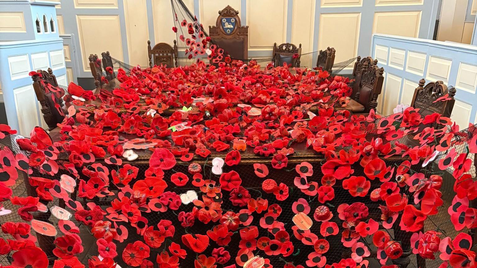 Red and black poppies made of fabric and paper spread over a large wooden table and chairs.