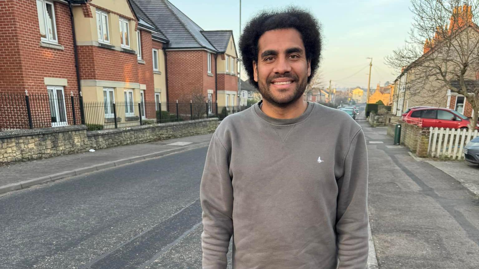 Sam Pearce-Kearney standing on a residential street. He is wearing a light grey crew-neck jumper. He has thick dark-brown afro hair and dark-brown stubble.