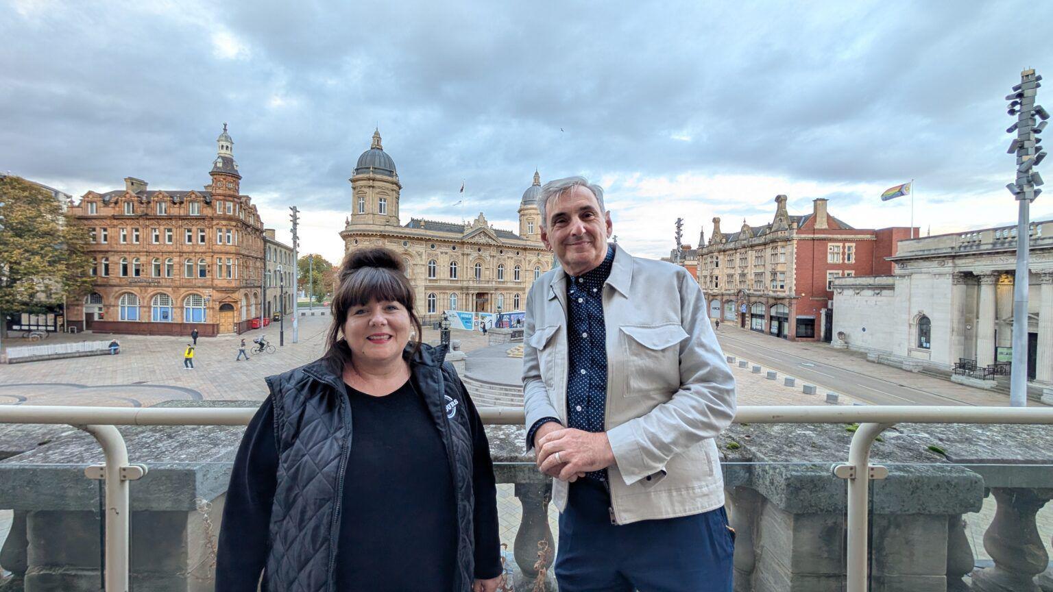 A woman with brown hair in a full fringe with a top bun is smiling into the camera and wearing a black body warmer and long top. Next to her is a tall man with grey hair, a white jacket, blue shirt and trousers. Behind them in Queen Victoria Square in Hull and they are stood on an elevated area.