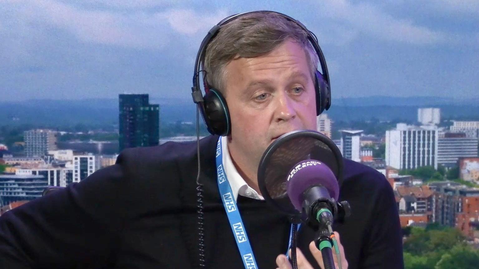 James Roach wearing a black jumper on top of a white shirt and a blue NHS lanyard. He is wearing headphones and his face is partially obscured by a microphone and pop filter. He is in the studio and the studio backdrop shows an image of Southampton's skyline. 