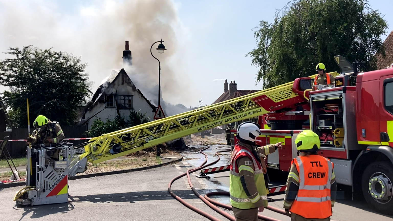 Smoke is coming out of the roof of a thatched cottage in the background. On the right, two firefighters are talking to each other while standing in front of a fire engine. Another firefighter is in the bucket of an aerial ladder. He has either just been returned to the ground or is about to be lifted up.