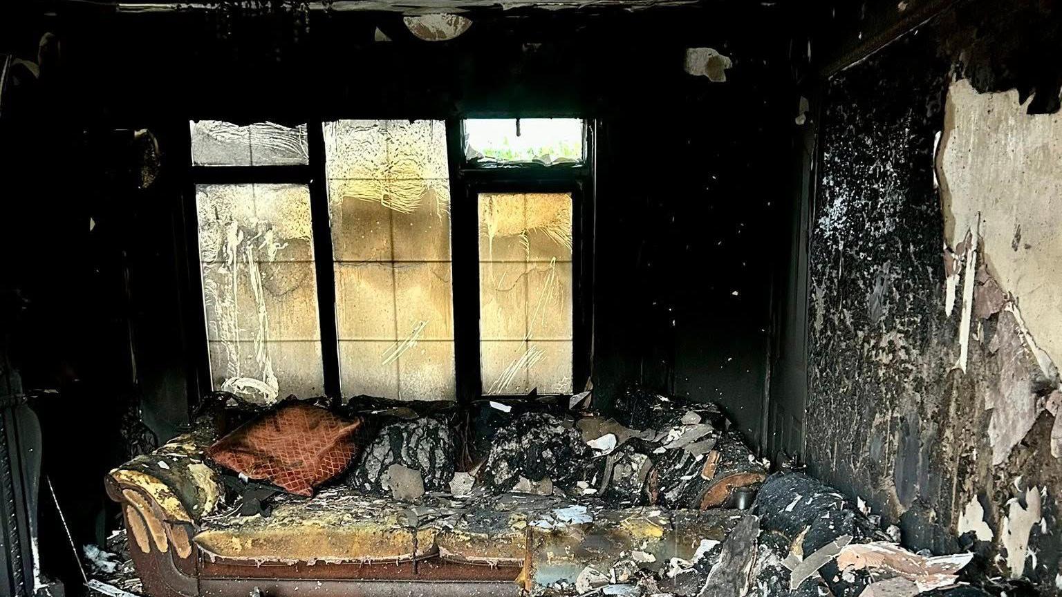 The inside of a living room which has been ravaged by fire. Huge chunks of paint and plaster are falling off the walls, the sofa is singed and the window is warped.