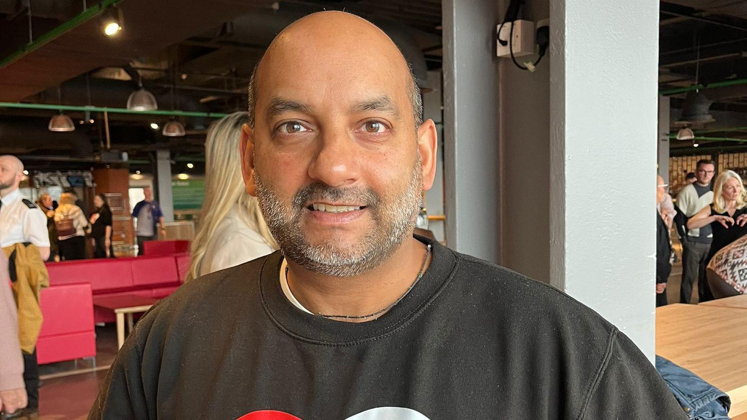 Nikhil Misra, who has a bald head and grey stubble, smiles as the camera wearing a black sweatshirt with a Knifesavers logo consisting of two hands forming the shape of a heart.