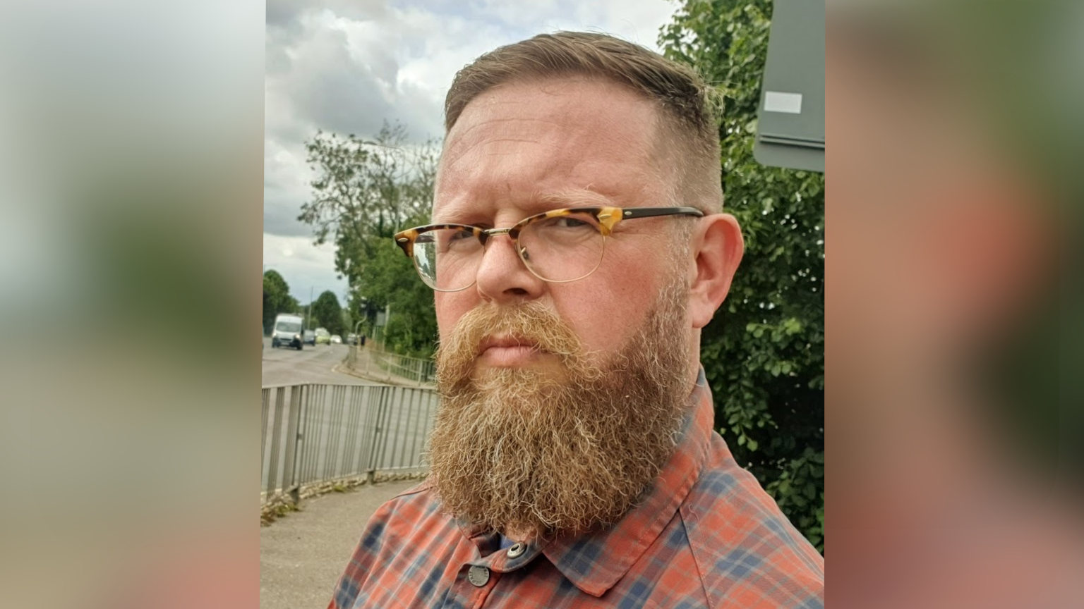 Liam Calvert, who has glasses and a beard, looks at the camera while standing on a street. He is wearing a red/orange and blue checked shirt