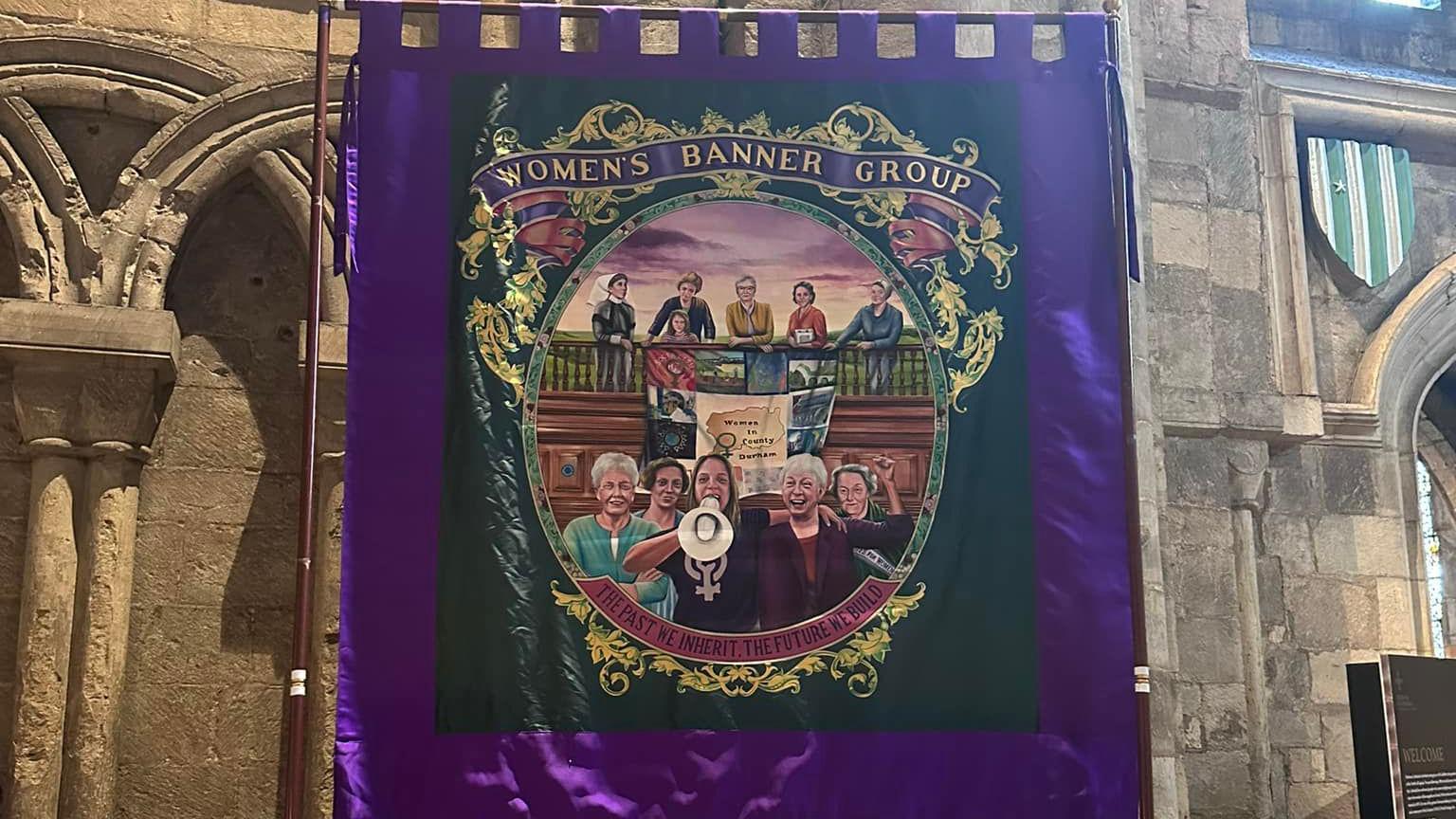 A purple and green silk banner with 11 women on, with one holding up a megaphone. At the top, it reads 'Women's Banner Group' on a purple ribbon. The banner is hanging on large poles and resting on the inside walls of a stone church.