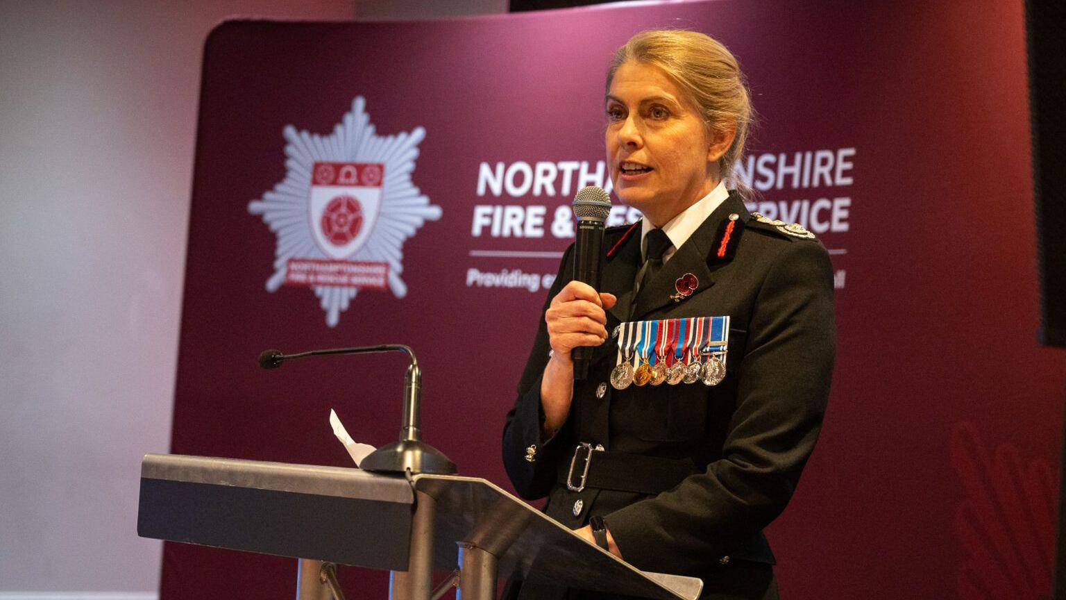 Nikki Watson, the chief fire officer, in a black, fire service uniform. There are medals pinned to her jacket. She is standing at a lectern and holding a microphone whilst making a speech. She is in front of a red, branded Northamptonshire Fire & Rescue Service banner.