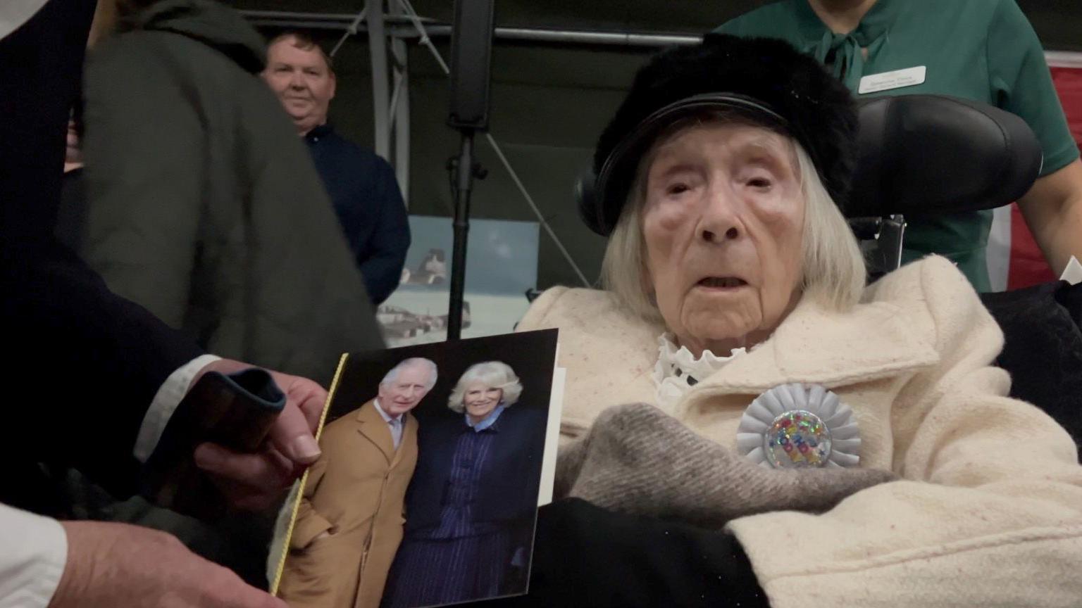 Dorothy Smith is sat in her wheelchair wearing a white coat and is being handed an A5 card. On the front of the card is a picture of the King and Queen.