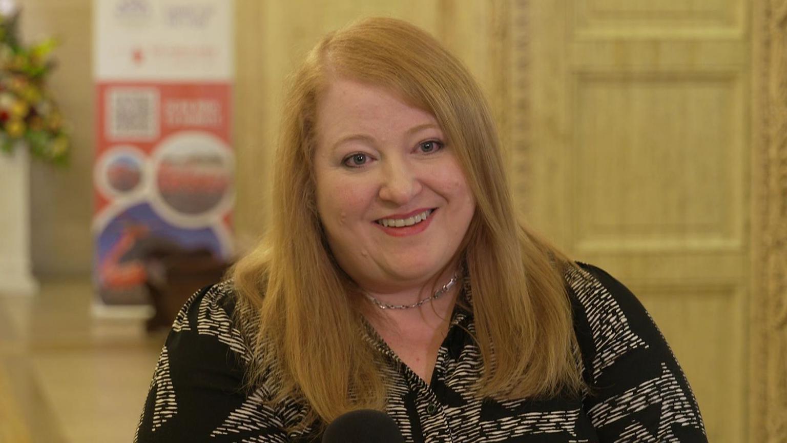 Naomi Long has red hair, worn down. She is smiling and has pink lipstick on. She wears a silver necklace, and a black and white shirt. 