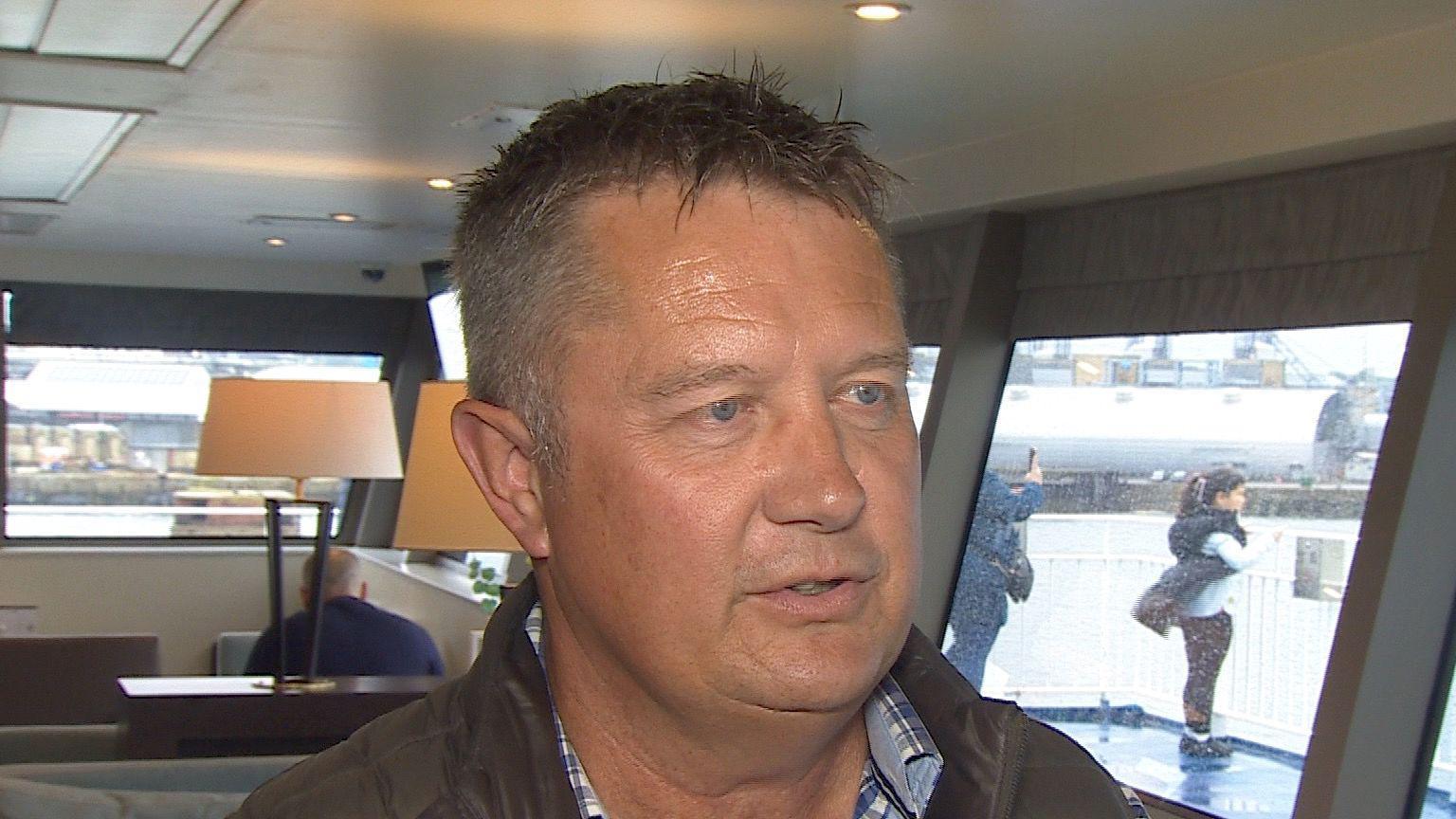 A white middle-aged man with short brown hair, with a ferry interior in the background