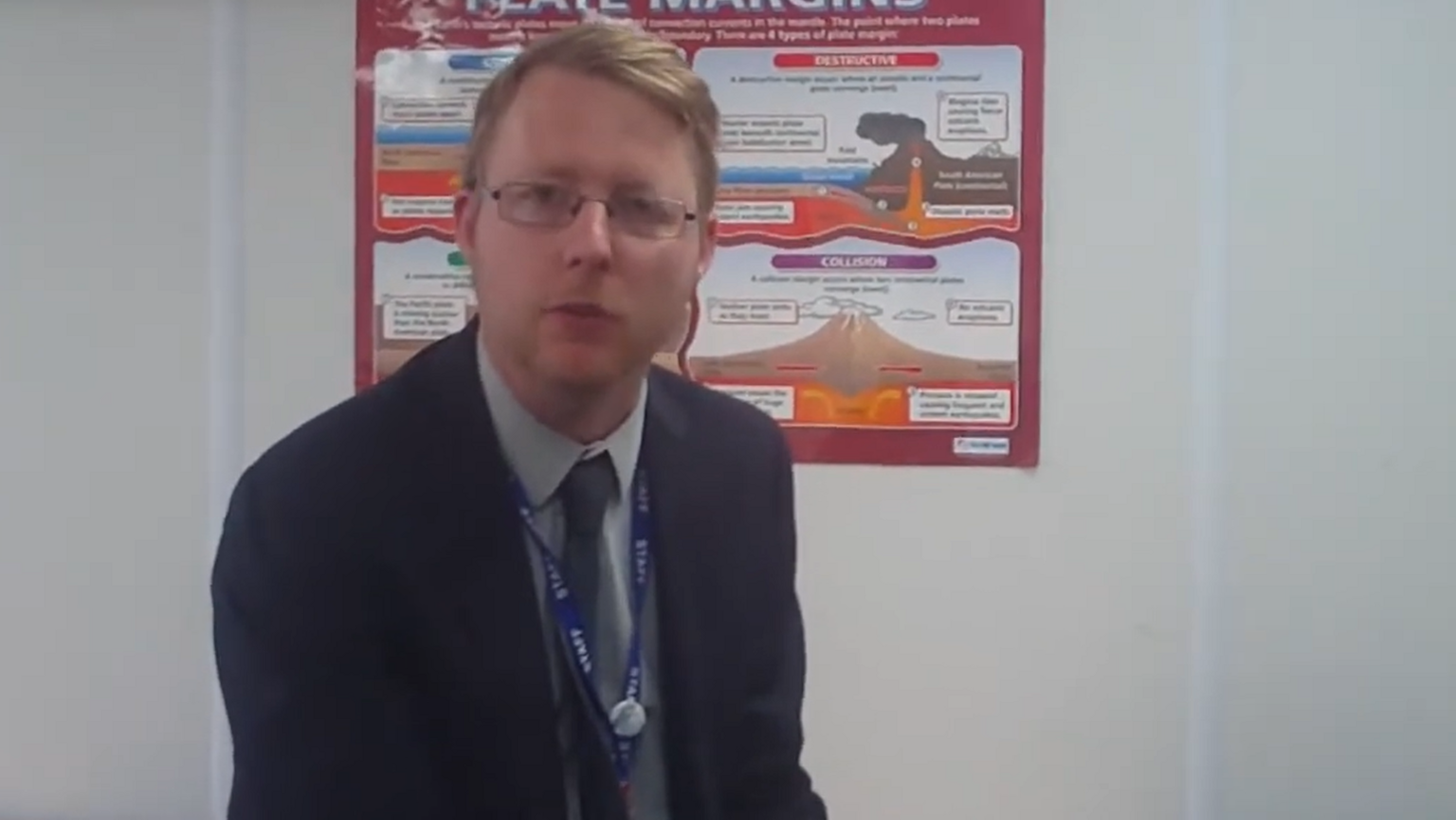 A screen-grab of a video which shows Sean Rainforth in a classroom wearing a blue staff lanyard. A poster behind him explains tectonic plates.