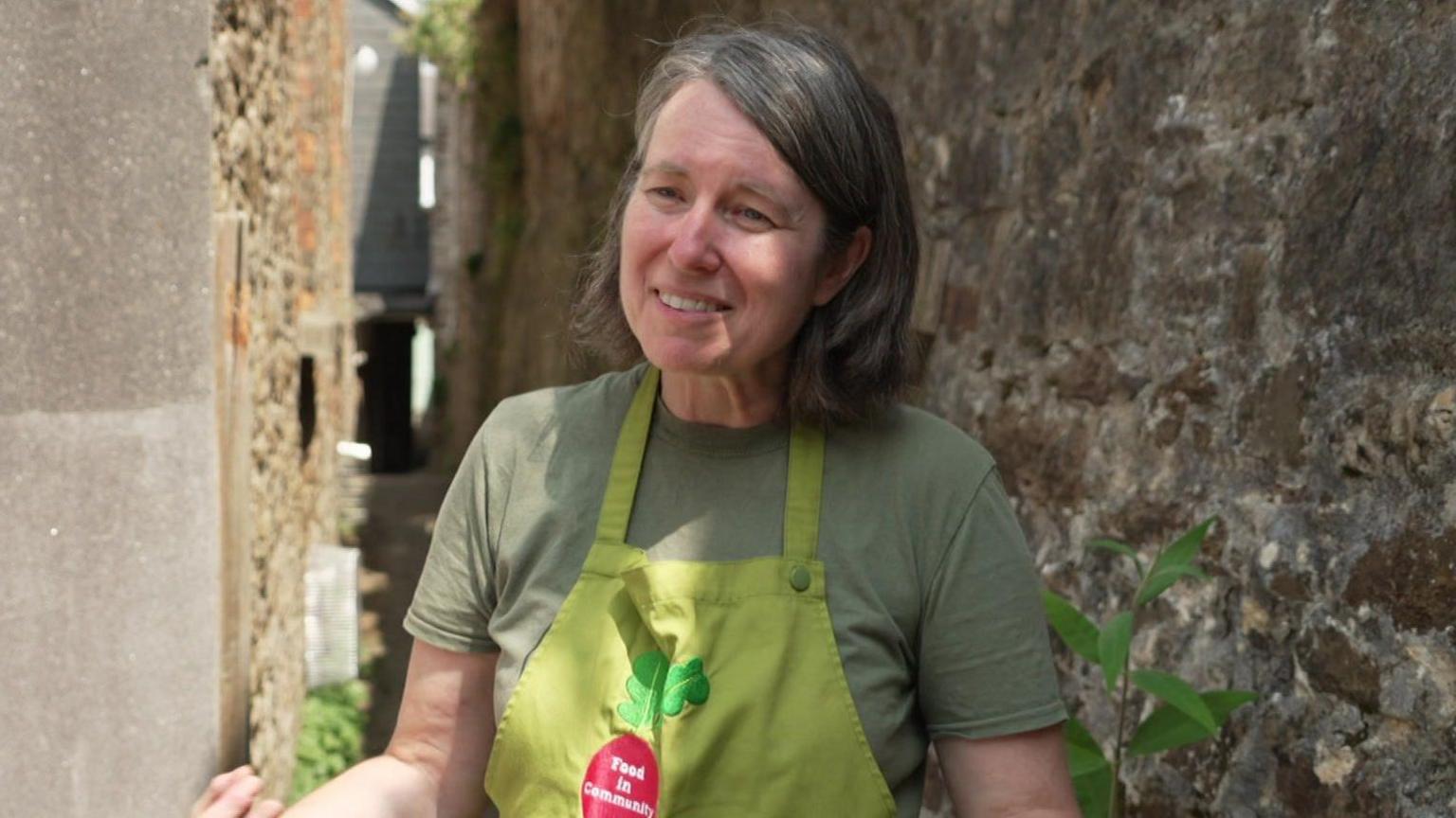 Chantelle Norton, a director of Food in Community CIC, stands smiling in front of a wall covered with lichen and moss. She is wearing an olive green apron bearing the words "Food in Community" over a khaki short sleeved T shirt. Her shoulder length salt and pepper hair is styled in a long bob.
