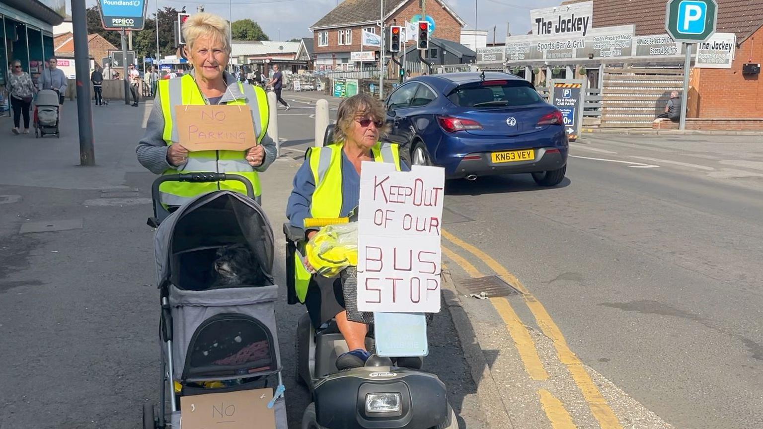 Two bus and mobility scooter users hold placards at the side of the road. One woman, with blonde hair and a Hi-Vis jacket on is holding a sign that reads 'no parking'. Another one, sitting on a mobility scooter, is wearing a blue T-shirt and a hi-vis jacket. She is holding a sign that reads 'keep out of our bus stop'. 