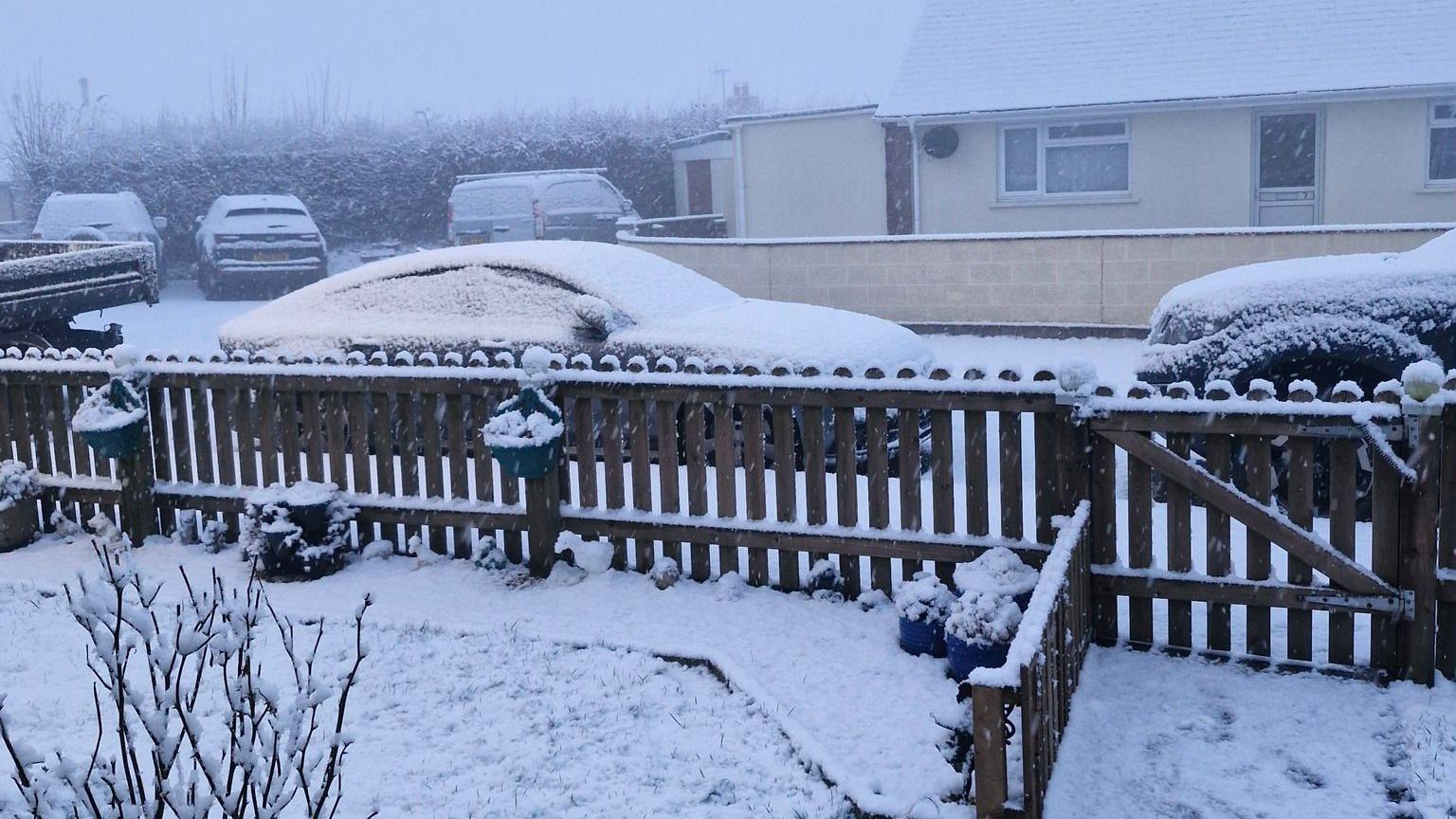 Five cars and vans parked on a residential street. Heavy snow has fallen and settled on their roofs and windscreens. There is a front garden fence in the foreground of the picture which is covered in snow. In the distance the sky is grey and foggy with low visibility. 