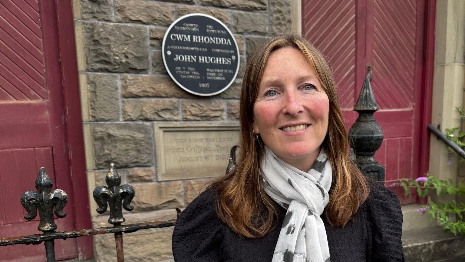 Rhian Hopkins outside the chapel in black top with white and black pattern scarf. She has long light brown hair and wears a silver hoop ear rings