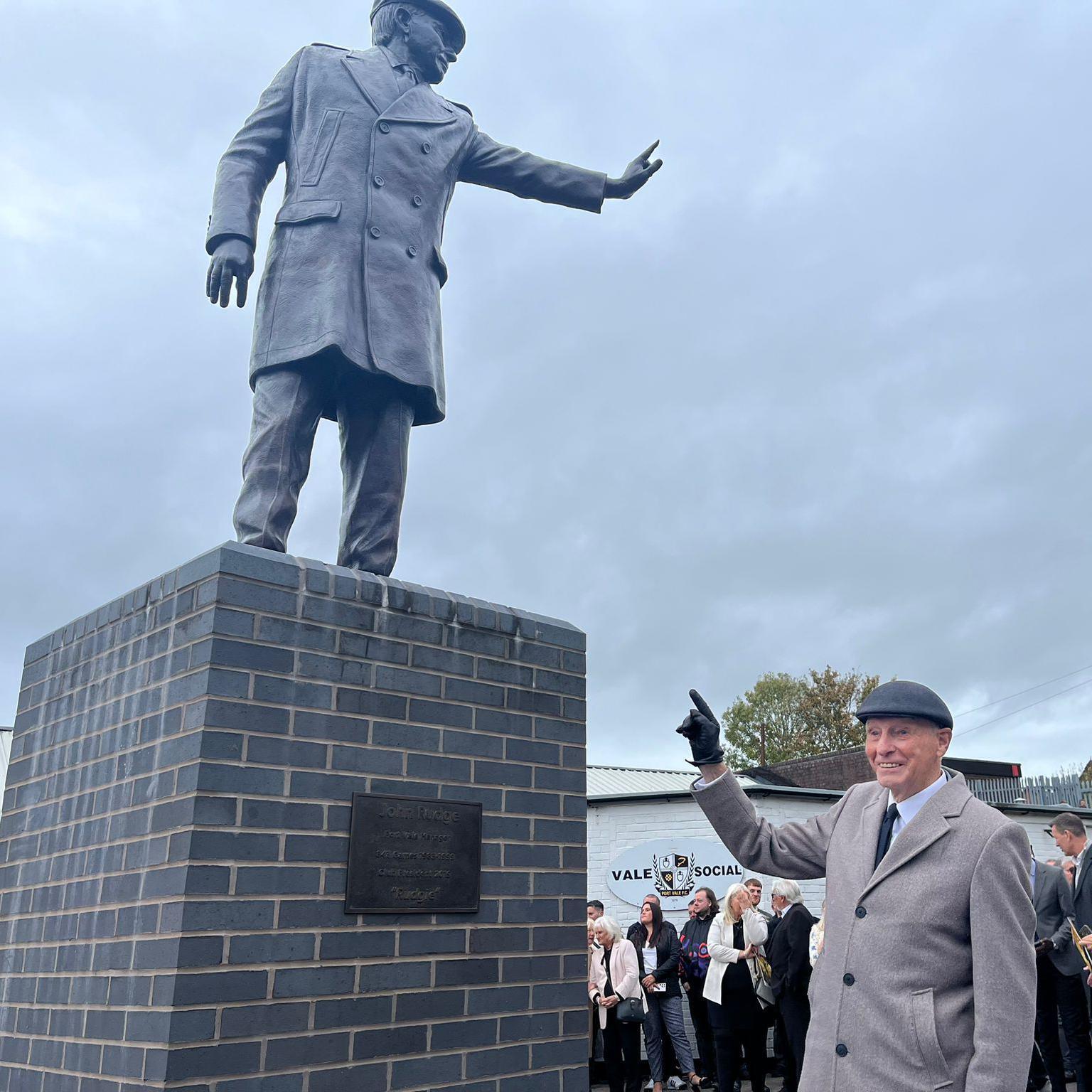 John Rudge, wearing a grey coat and flat cap smiles while pointing up to the statue of himself.