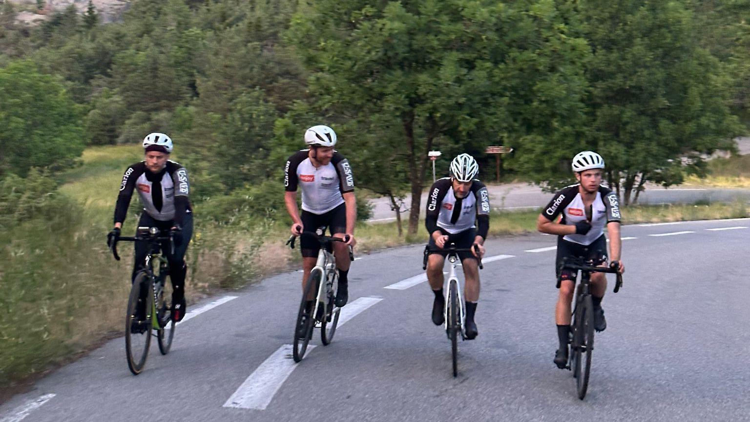 Four cyclists riding up a hill in the Alps, wearing red and white jackets and black shorts or trousers.